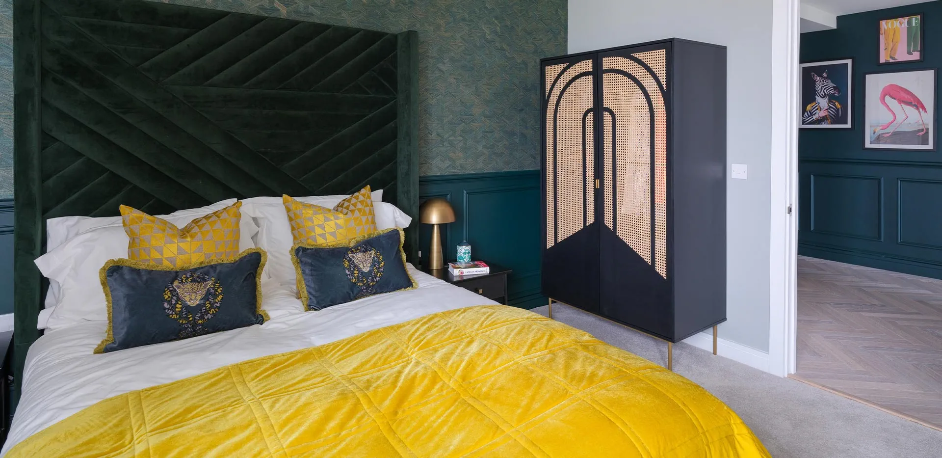 Stylish bedroom interior with dark green velvet headboard, yellow throw, and modern rattan wardrobe.